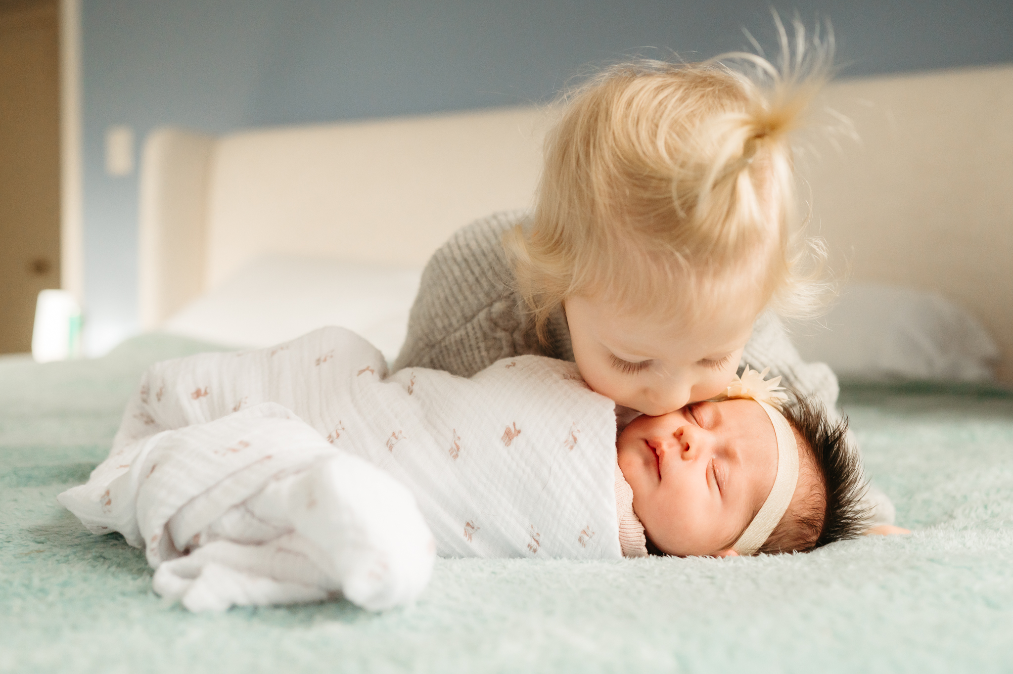 A cozy in-home newborn photography session featuring a sleeping baby wrapped in a soft blanket with an older sibling leaning over to kiss her, showcasing a natural and relaxed family environment