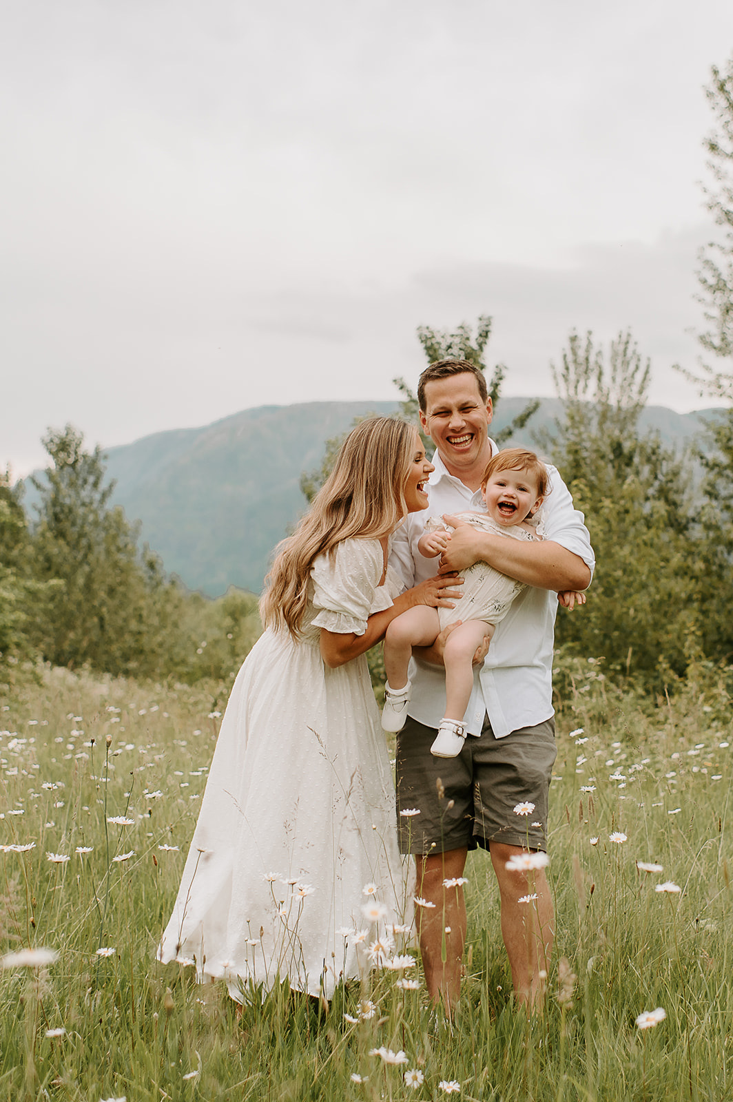 Joyful family of three laughing in a wildflower field with Mount Hood in the background during a spring family photo session in Portland. Mom in a flowing dress, dad in casual neutrals.