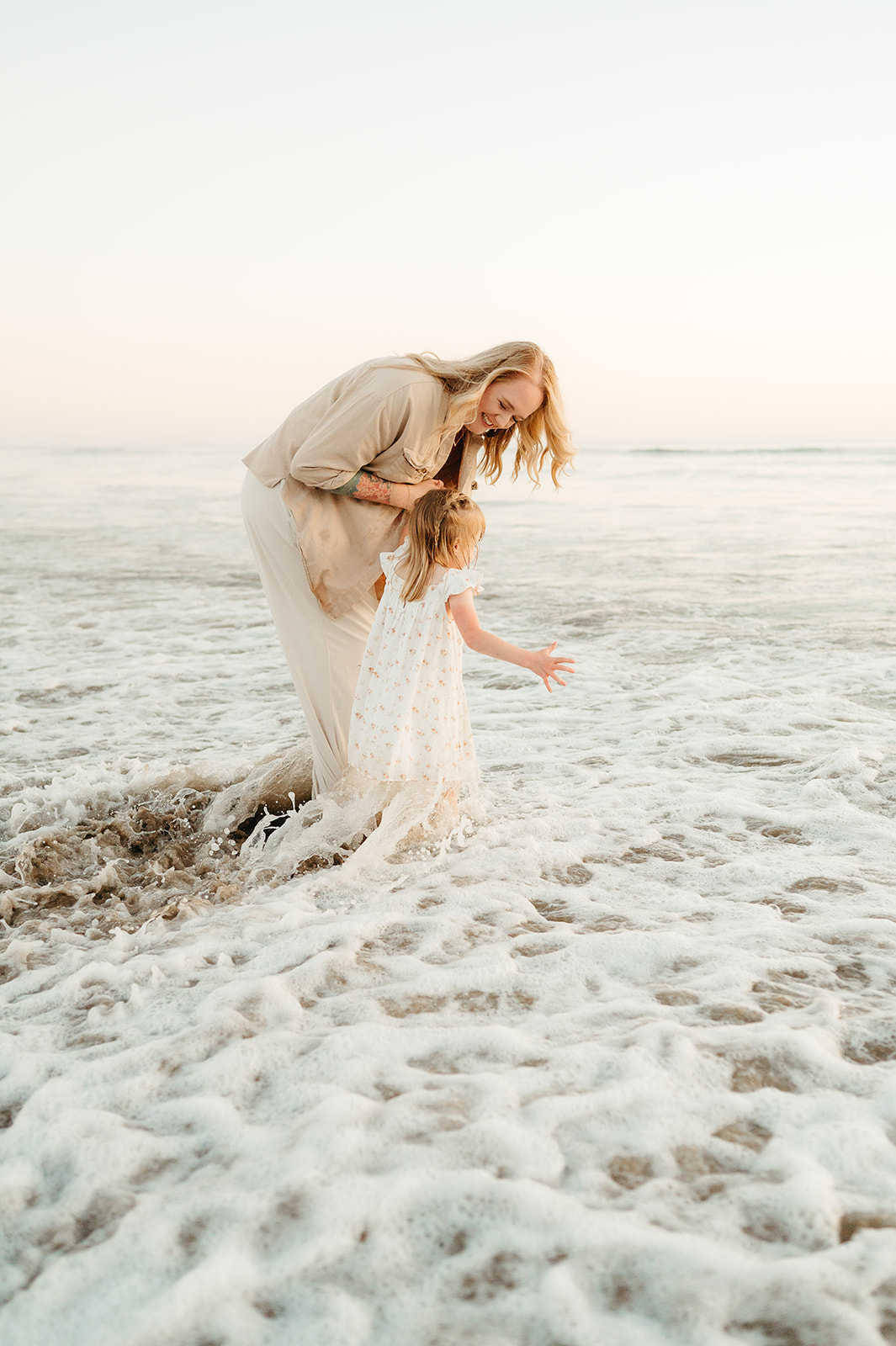 Mother and daughter play in the ocean waves during a sunset beach photography session on the Oregon coast. The mother, dressed in soft neutrals, laughs as her barefoot child splashes in the water, capturing a natural and carefree family moment