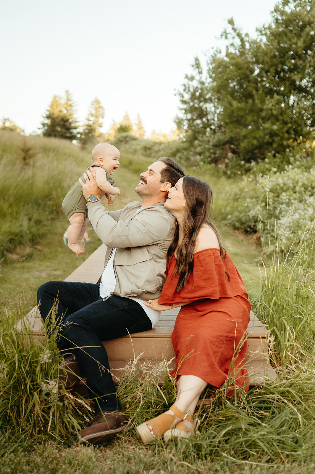 Parents sit on a wooden bench in a green meadow, smiling as they lift their baby into the air. A joyful, nature-filled family photography session with soft golden hour light.