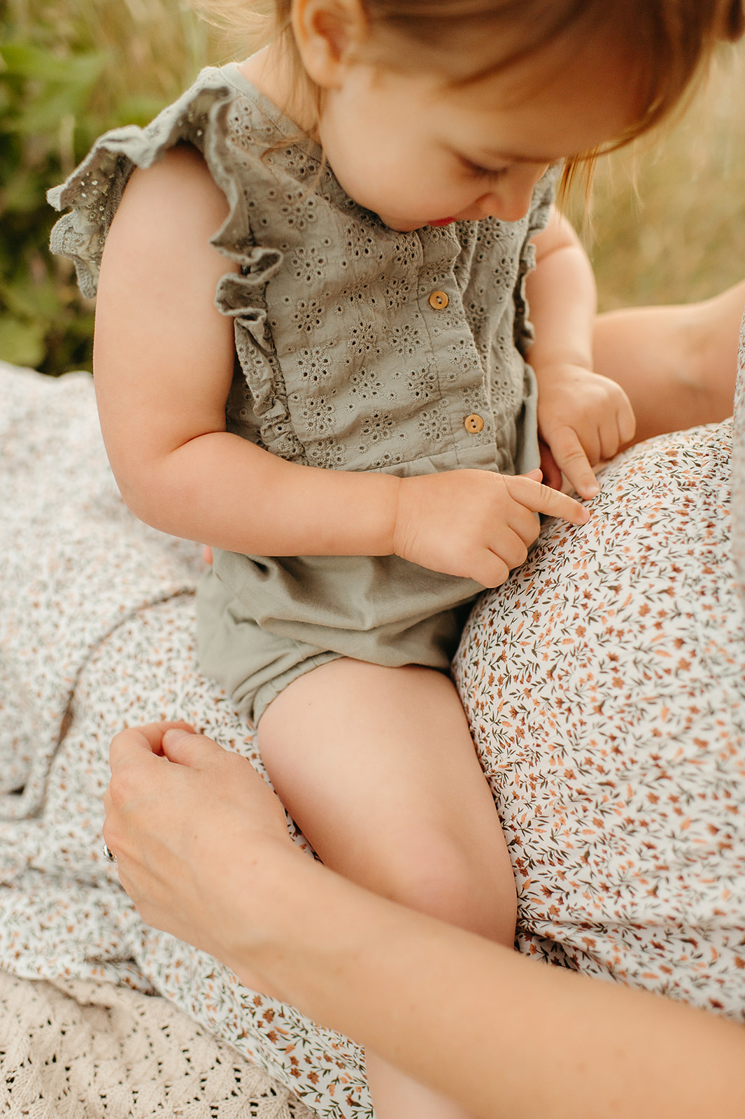 Close-up of a toddler sitting on their pregnant mother's lap, pointing at her baby bump. A sweet, intimate moment during a maternity session in a sunlit field, featuring soft floral textures and earthy tones.