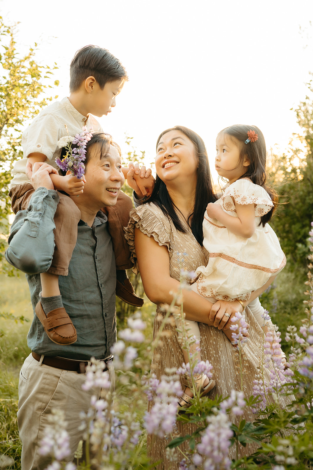 A family of four surrounded by blooming wildflowers, sharing a joyful moment during a golden hour spring session. Parents hold their children close, dressed in soft earth tones and neutral textures.