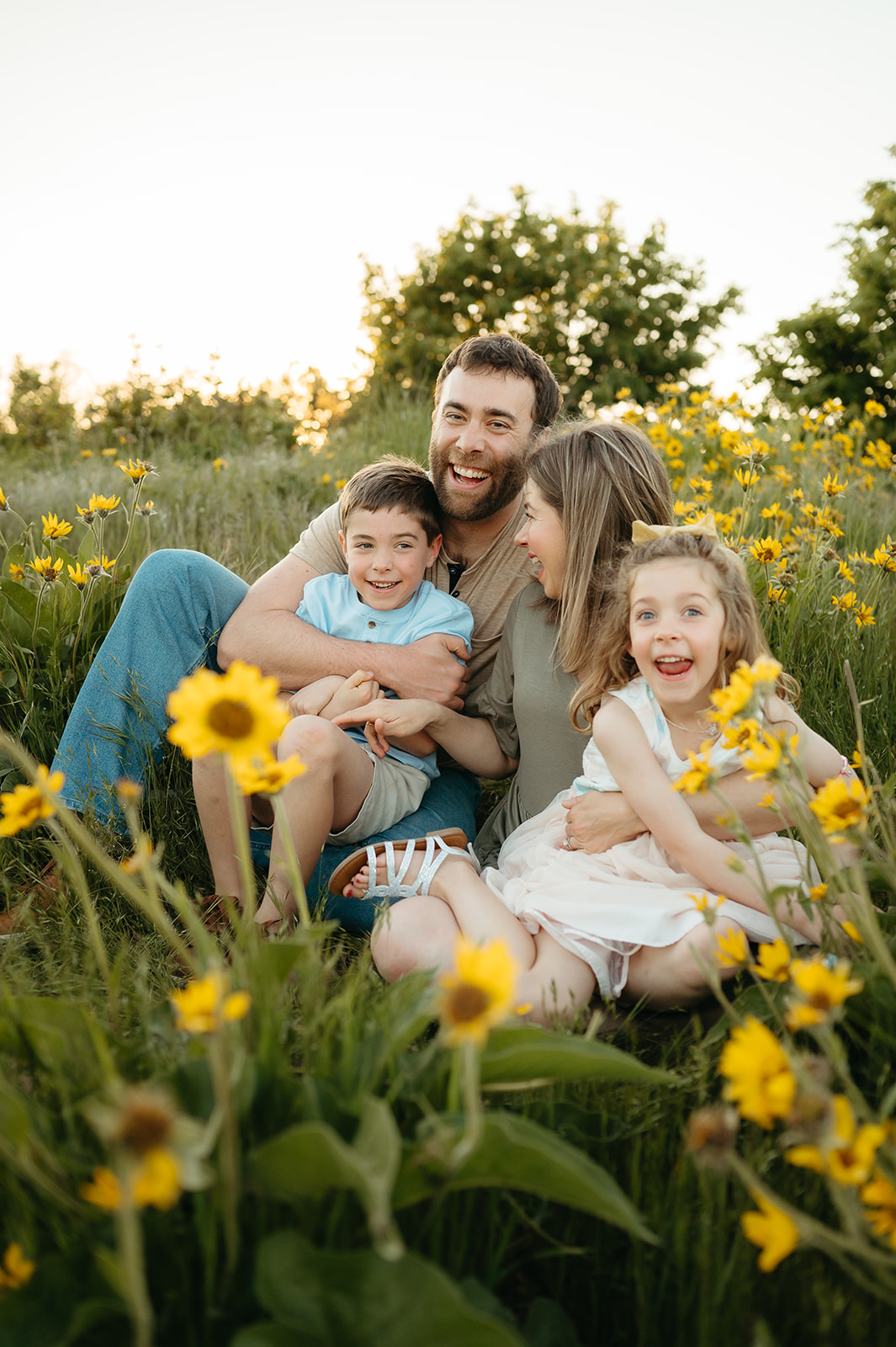 Family of four laughing together in a wildflower field at sunset. Parents embrace their children, capturing a warm and playful moment during a Portland family photo session.