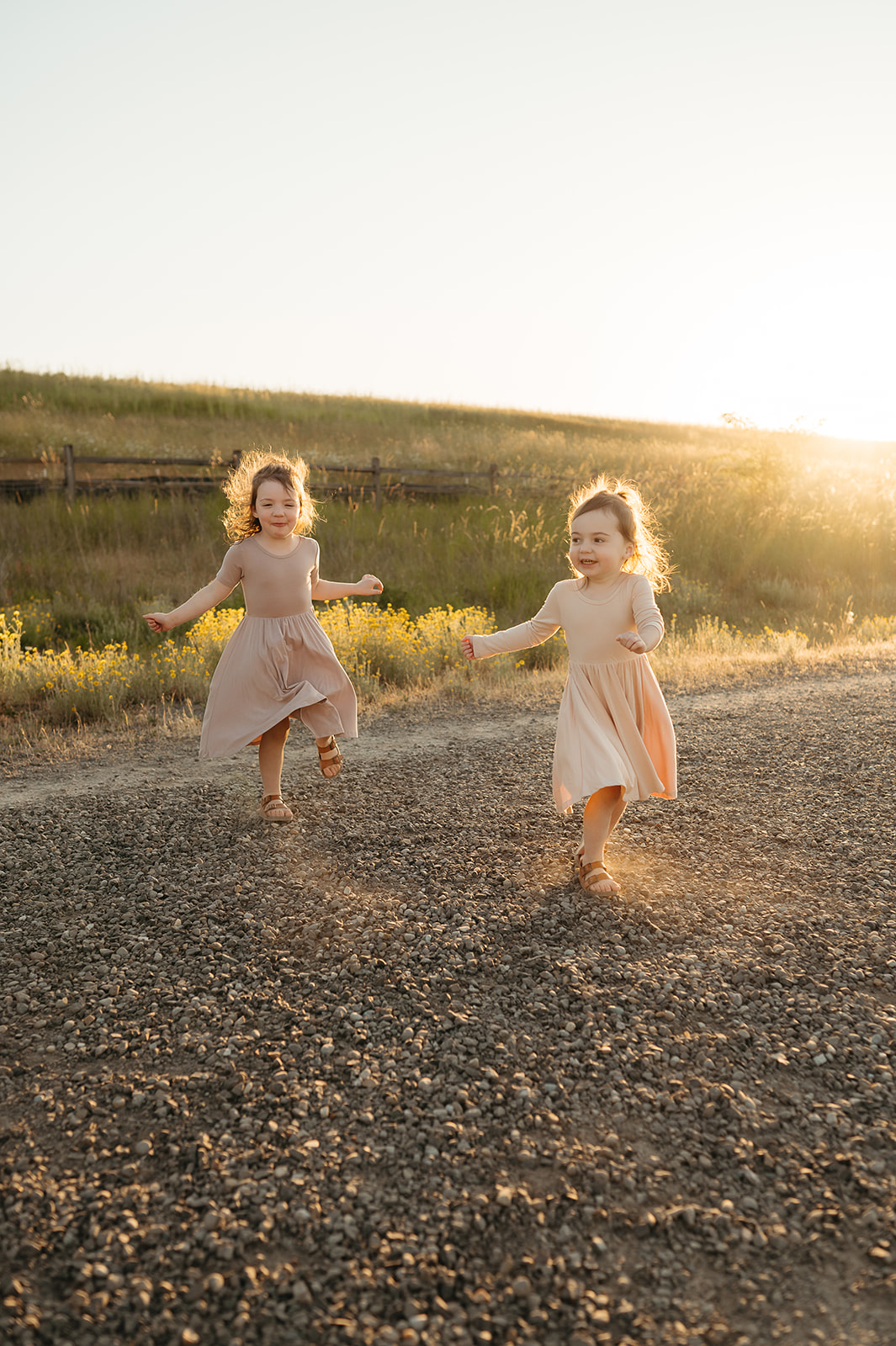 Two young sisters running barefoot on a gravel path at sunset, their dresses flowing in the golden light. A playful and carefree family photography moment Portland nature.