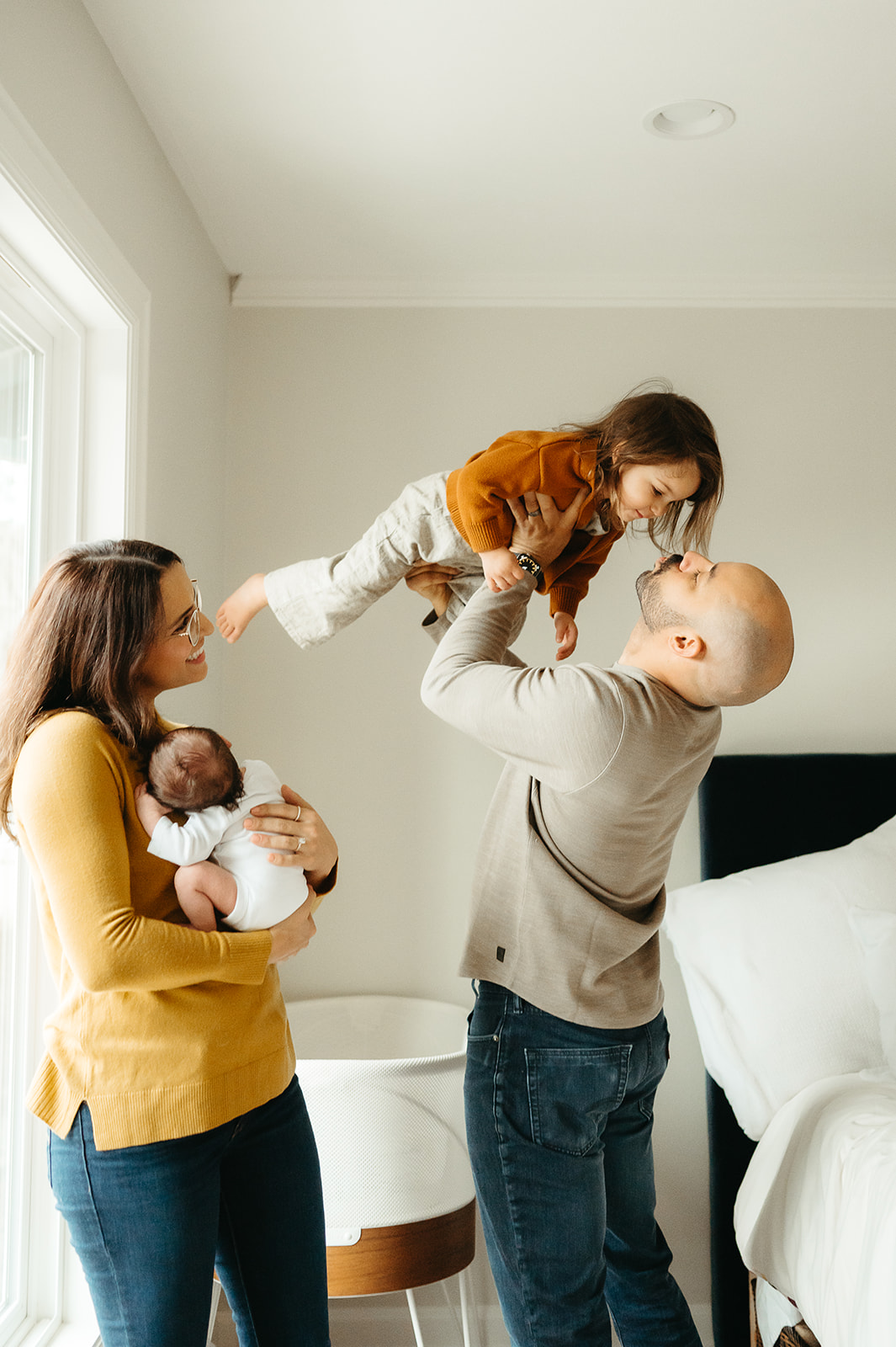 In-home Portland newborn session — mom holding baby by the window while dad lifts and kisses toddler on the bed, cozy and natural light lifestyle photo.