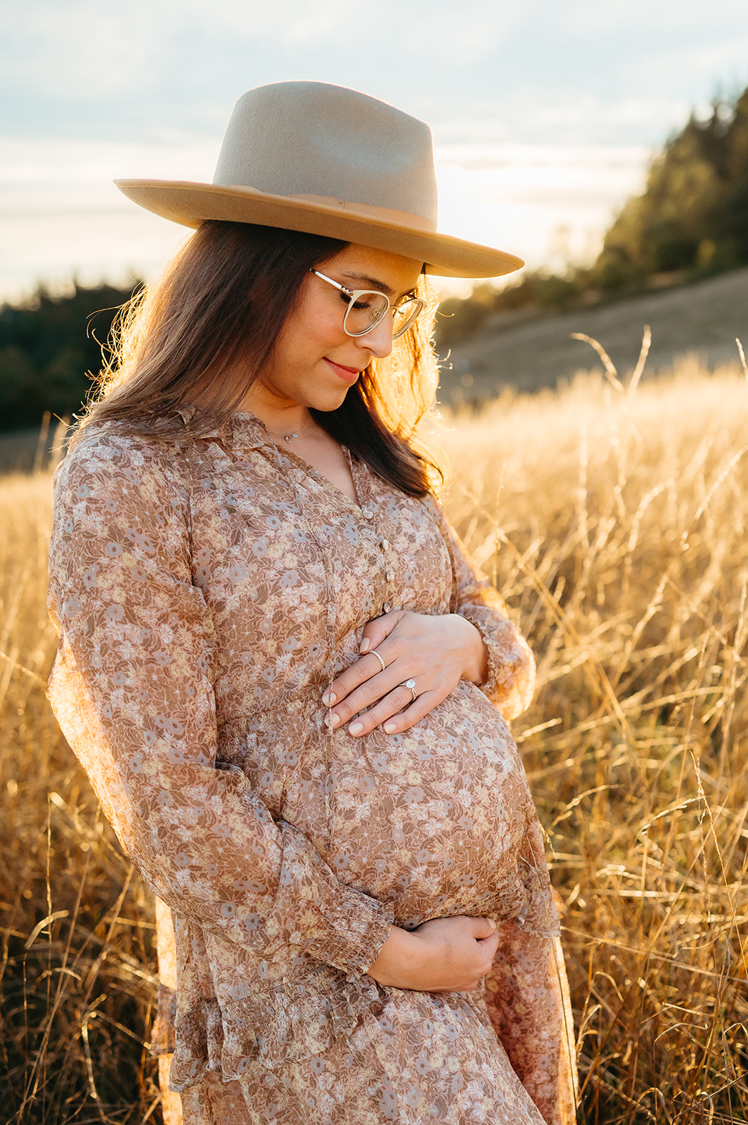 Pregnant mom wearing hat and floral dress looking down at belly in golden light field — Portland maternity photo.