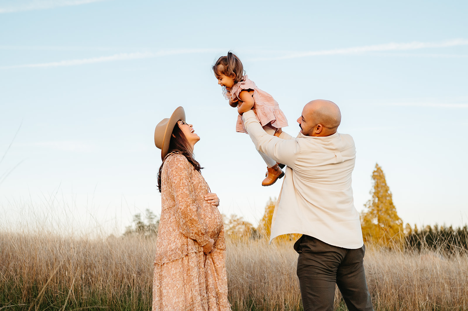 Portland maternity photo — pregnant mom holds belly while dad lifts toddler to kiss her at sunset in a golden field.