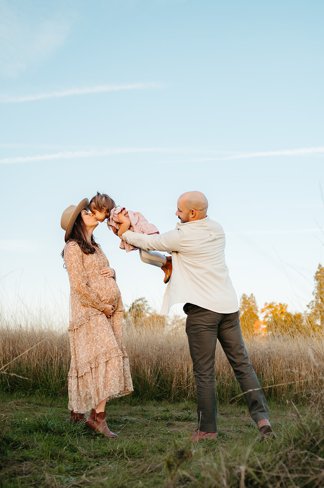 Outdoor Portland maternity photo — pregnant mom in floral dress holds bump while dad lifts toddler to kiss her at sunset in a golden field.